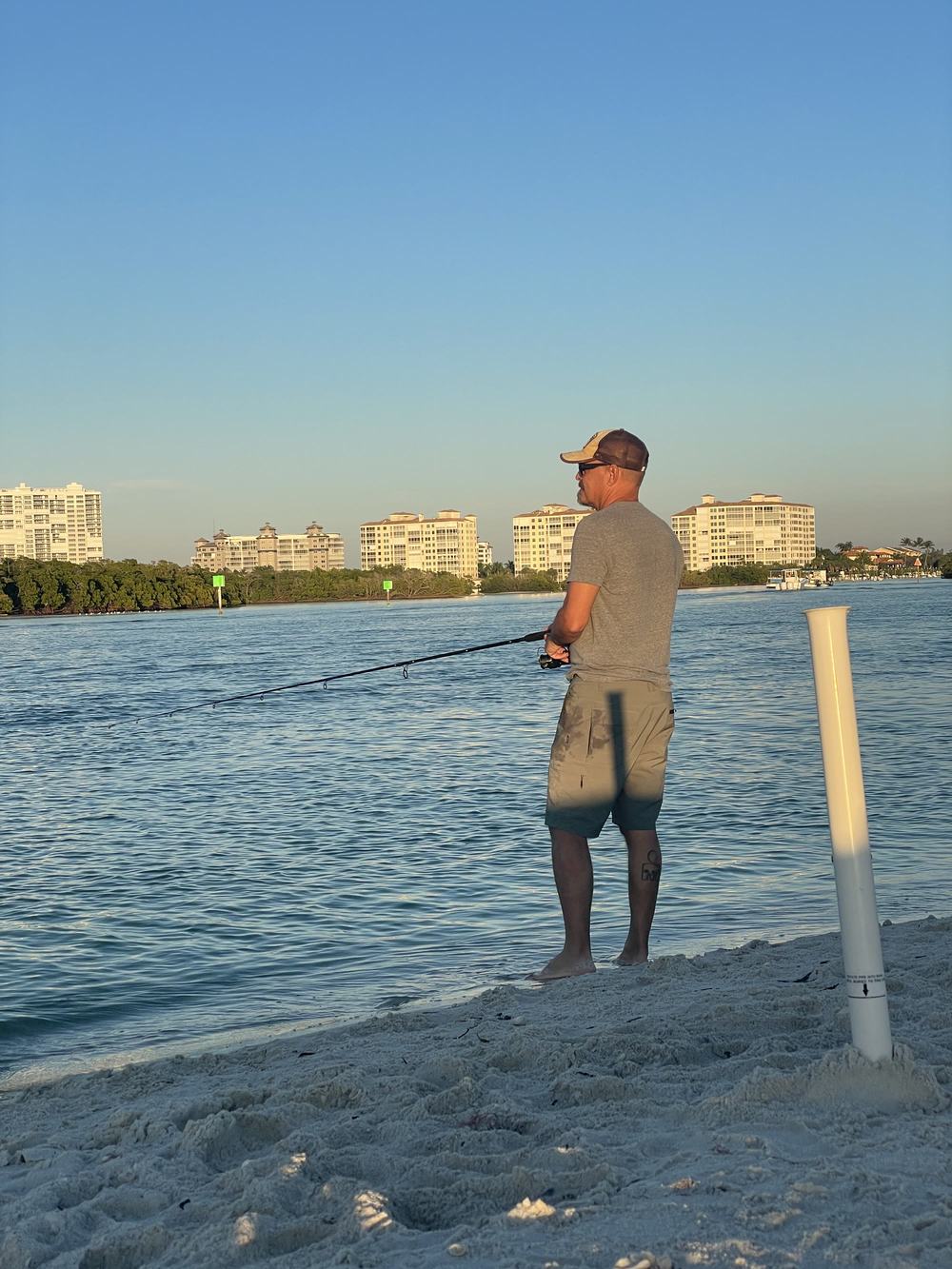 A person standing on a sandy beach, casting a fishing rod toward water with buildings on the far shore.
