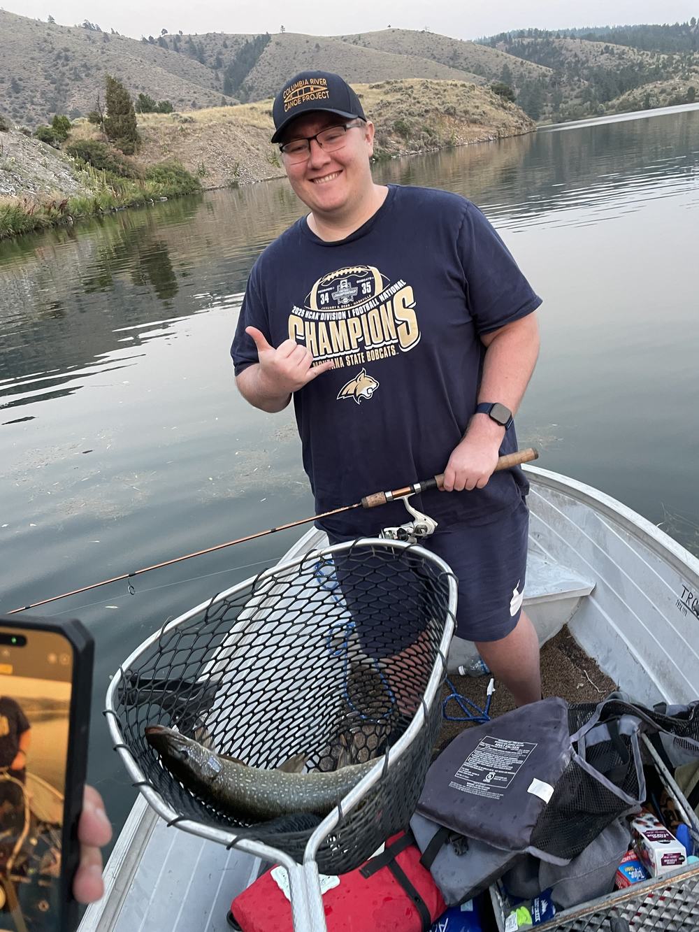 A person in a small boat holds a landing net with a pike, with grassy hills behind.