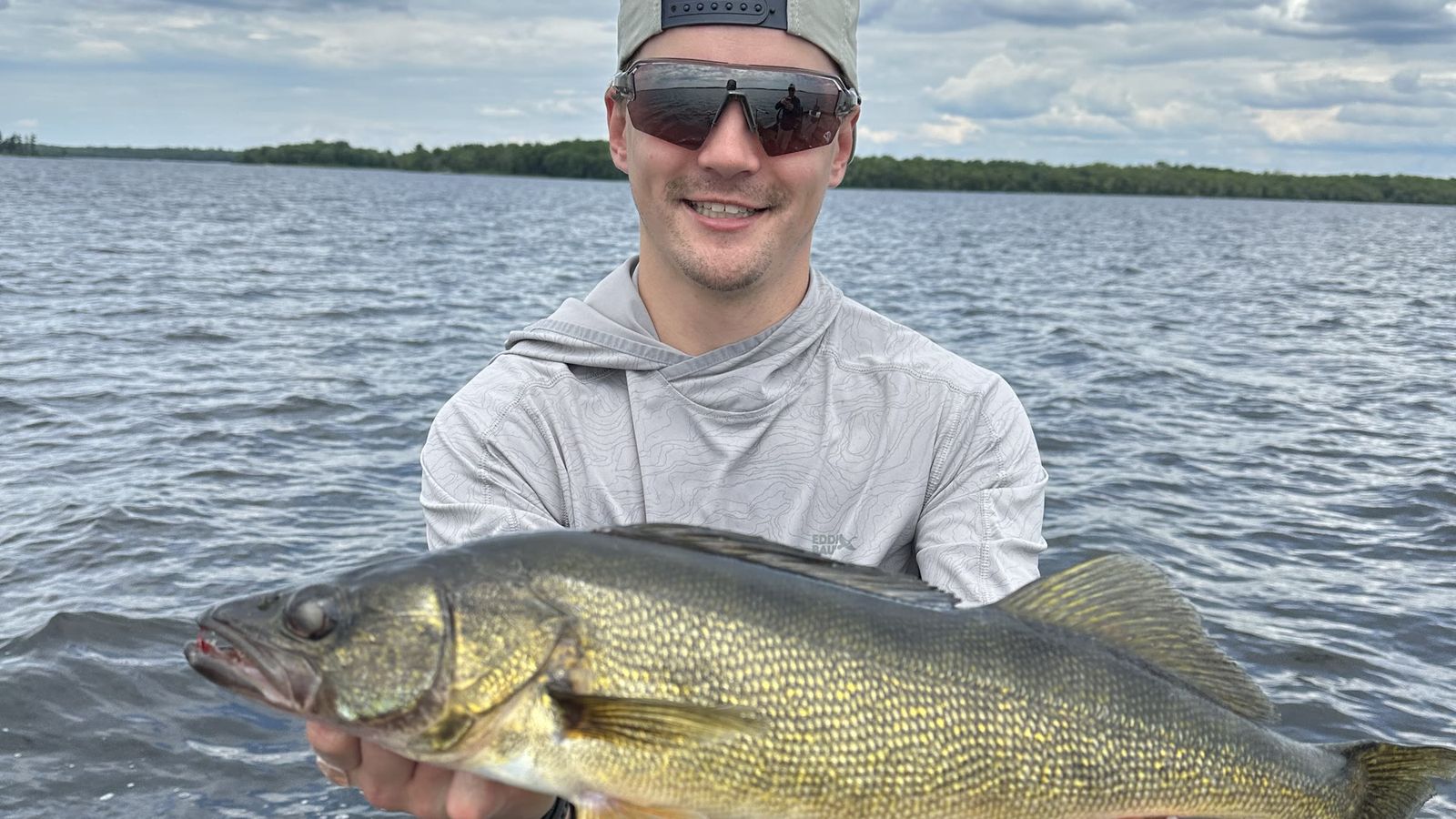 A walleye held over open water on a cloudy day, with a tree-lined shore behind.