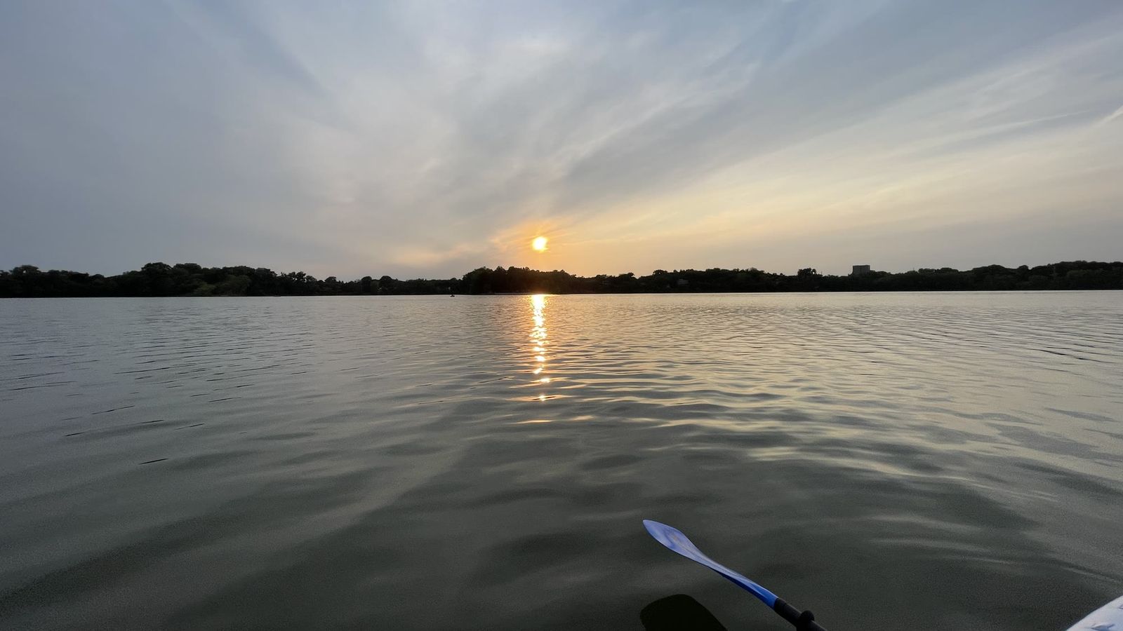 Sunset over a calm lake, seen from a paddleboard, with a paddle blade in the foreground.